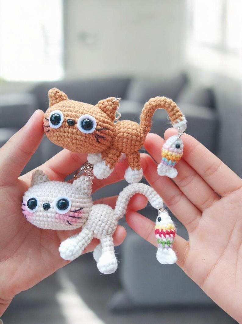 May include: Two crocheted cat keyrings. One is tan with black stripes and white paws, the other is white with pink cheeks. Each cat has large black eyes and a tail with a small, colourful fish attached.