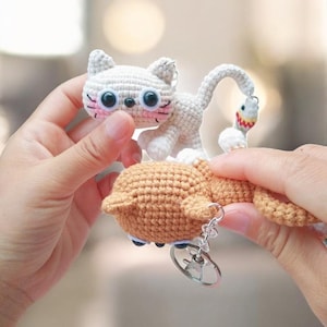 May include: A crocheted cat keyring featuring a light grey cat and a brown fish. The cat has large black eyes, pink cheeks, and a small black nose. The fish has white body with red and yellow details. The keyring is attached to a silver ring.