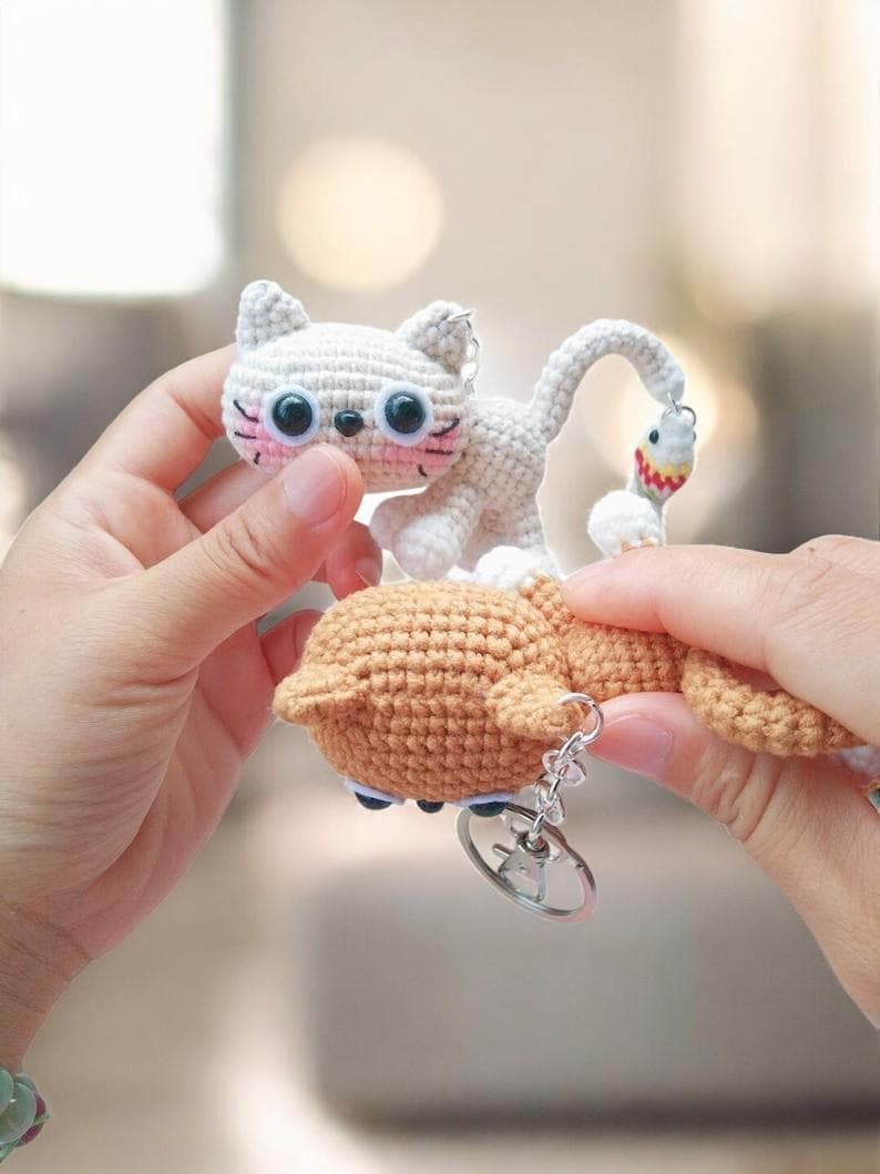 May include: A crocheted cat keyring featuring a light grey cat and a brown fish. The cat has large black eyes, pink cheeks, and a small black nose. The fish has white body with red and yellow details. The keyring is attached to a silver ring.