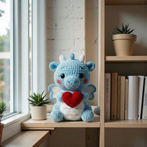 May include: A light blue crocheted dragon plushie with a red heart, white wings, and white horns. The dragon is sitting on a wooden shelf near a window. A small potted succulent is to the left of the dragon.