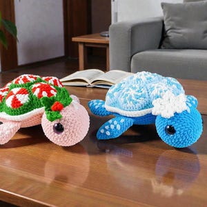 May include: Two handmade crochet turtle plushies. One is pink with a green and red shell design, the other is blue with white accents. Both have large, black eyes and are displayed on a wooden table.