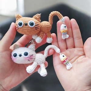 May include: Two crochet cat keyrings, one tan and one white, each with a dangling fish charm. The tan cat has white paws and large blue eyes. The white cat has pink cheeks and blue eyes. The fish charms have colourful stripes.