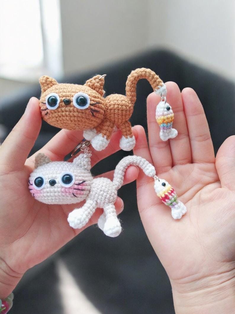 May include: Two crochet cat keyrings, one tan and one white, each with a dangling fish charm. The tan cat has white paws and large blue eyes. The white cat has pink cheeks and blue eyes. The fish charms have colourful stripes.