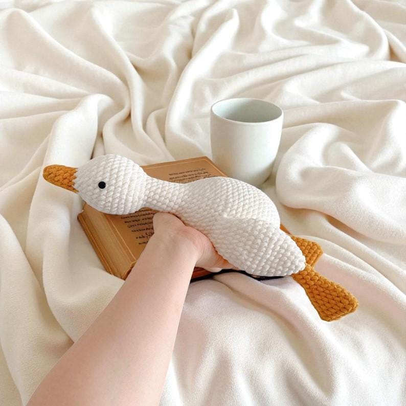 May include: A white crocheted goose toy with a textured surface, featuring a bright orange beak and orange feet. The goose has a small black eye and is crafted with a visible yarn stitch pattern, presenting a soft and rounded form throughout its body.