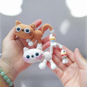 May include: Two crocheted cat keyrings. One is orange with white paws, the other is white with pink cheeks. The orange cat has a long tail and a small fish charm. The white cat has a fish charm as well. Both have large black eyes.