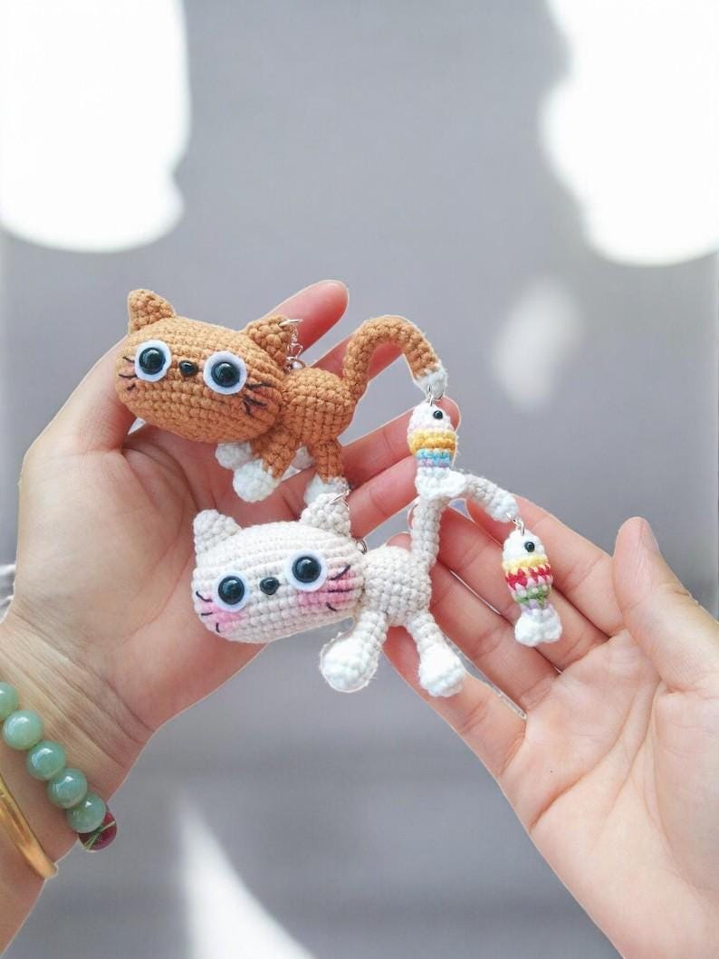 May include: Two crocheted cat keyrings. One is orange with white paws, the other is white with pink cheeks. The orange cat has a long tail and a small fish charm. The white cat has a fish charm as well. Both have large black eyes.