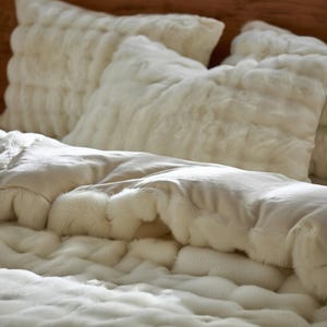 Könnte beinhalten: Nahaufnahme eines Bettes mit cremefarbenen Kunstfellkissen und einer passenden Decke. Die Kissen sind rechteckig und flauschig, und die Decke hat ein weiches, strukturiertes Aussehen. Die Bettwäsche schafft eine gemütliche und einladende Atmosphäre.