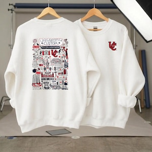 Puede incluir: Dos sudaderas blancas. Una presenta un gr&aacute;fico detallado con el texto "UNIVERSITY CUSTOM" y varias ilustraciones en rojo, negro y blanco. La otra tiene un logotipo "UC" rojo. Ambas tienen mangas largas y cuello redondo.