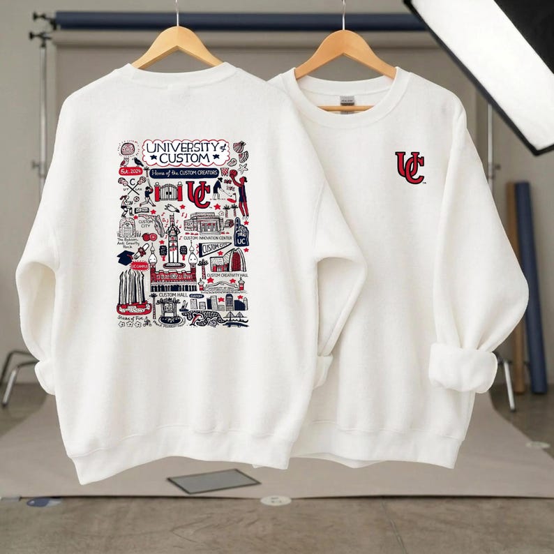 Puede incluir: Dos sudaderas blancas. Una presenta un gr&aacute;fico detallado con el texto "UNIVERSITY CUSTOM" y varias ilustraciones en rojo, negro y blanco. La otra tiene un logotipo "UC" rojo. Ambas tienen mangas largas y cuello redondo.