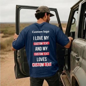 May include: A navy blue short-sleeved t-shirt featuring white and red text on the back that reads: Custom logo, I LOVE MY CUSTOM TEXT AND MY CUSTOM TEXT LOVES CUSTOM TEXT. The shirt is worn with a grey baseball cap and dark denim jeans.