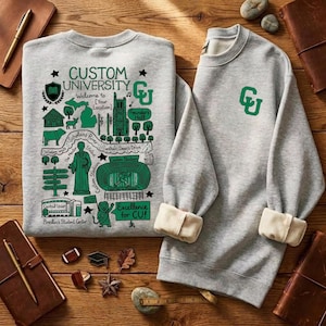 Puede incluir: Dos sudaderas grises con pu&ntilde;os color crema. Una presenta un gr&aacute;fico verde con el texto "CUSTOM UNIVERSITY" y un dise&ntilde;o de mapa. La otra tiene un logotipo "CU" verde. Sobre una superficie de madera.
