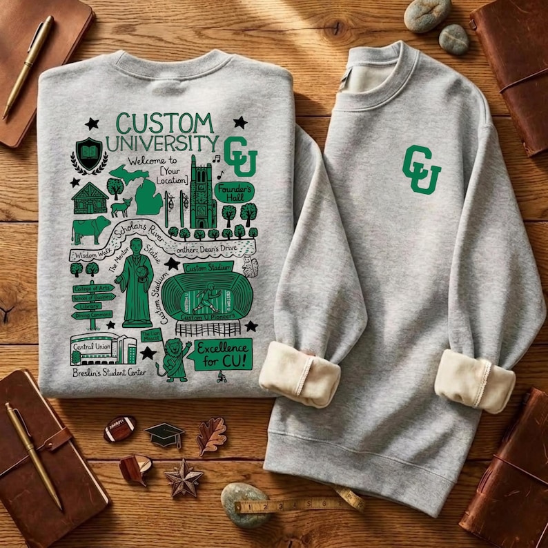 Puede incluir: Dos sudaderas grises con pu&ntilde;os color crema. Una presenta un gr&aacute;fico verde con el texto "CUSTOM UNIVERSITY" y un dise&ntilde;o de mapa. La otra tiene un logotipo "CU" verde. Sobre una superficie de madera.