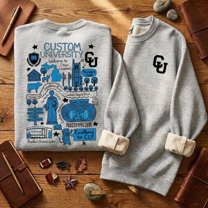 Puede incluir: Dos sudaderas grises con pu&ntilde;os color crema se muestran sobre una superficie de madera. Una sudadera presenta un gr&aacute;fico azul con el texto "CUSTOM UNIVERSITY" y varias ilustraciones. La otra tiene un logotipo "CU" negro.