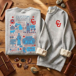 Puede incluir: Dos sudaderas grises con pu&ntilde;os color crema y cuello redondo se muestran sobre una superficie de madera. Una sudadera presenta un gr&aacute;fico detallado en azul y rojo con el texto "CUSTOM UNIVERSITY" y "Excellence for CU!". La otra tiene un logotipo "CU" rojo.