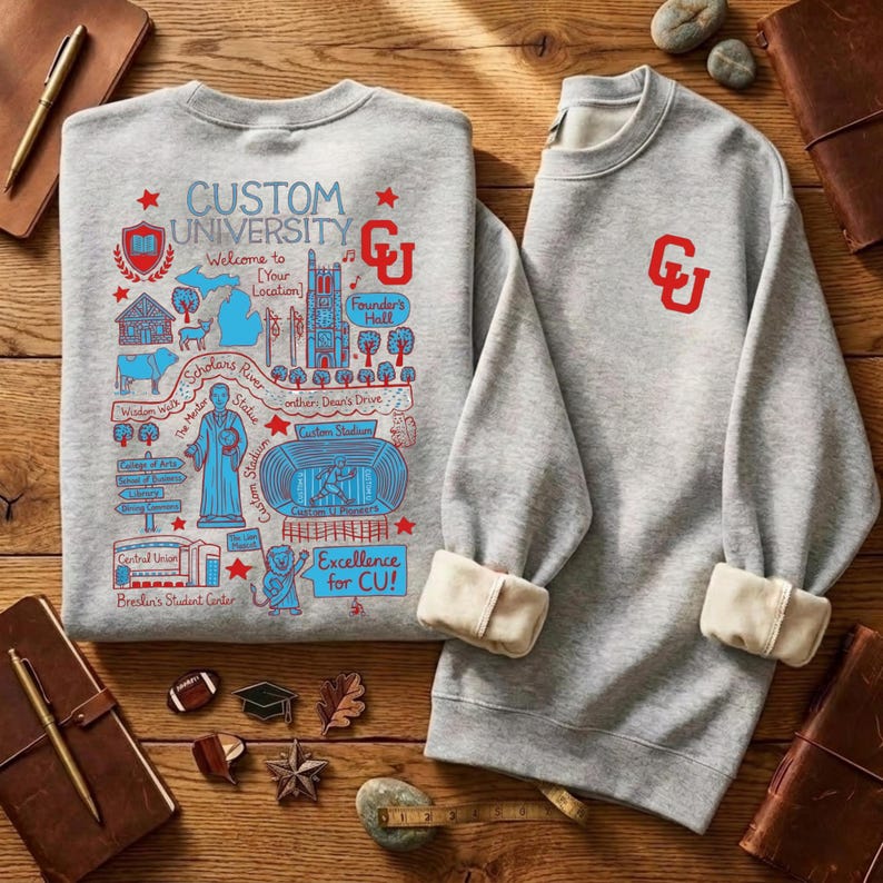 Puede incluir: Dos sudaderas grises con pu&ntilde;os color crema y cuello redondo se muestran sobre una superficie de madera. Una sudadera presenta un gr&aacute;fico detallado en azul y rojo con el texto "CUSTOM UNIVERSITY" y "Excellence for CU!". La otra tiene un logotipo "CU" rojo.