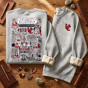 Puede incluir: Dos sudaderas grises. Una presenta un gr&aacute;fico detallado con el texto "UNIVERSITY of CUSTOM" y "Home of the CUSTOM CREATORS". La otra tiene un logotipo "UC" rojo. Los pu&ntilde;os est&aacute;n doblados.