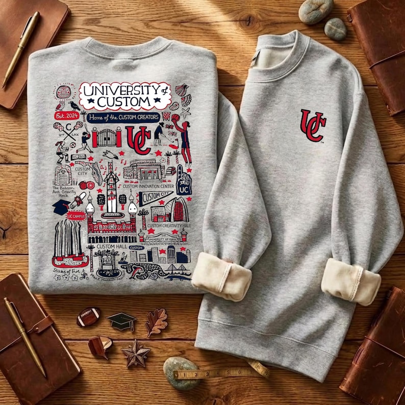 Puede incluir: Dos sudaderas grises. Una presenta un gr&aacute;fico detallado con el texto "UNIVERSITY of CUSTOM" y "Home of the CUSTOM CREATORS". La otra tiene un logotipo "UC" rojo. Los pu&ntilde;os est&aacute;n doblados.