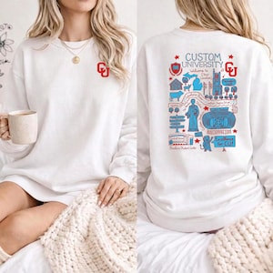 Puede incluir: Sudadera blanca con el logo "CU" rojo. La parte trasera presenta un gr&aacute;fico detallado en rojo y azul con el texto "CUSTOM UNIVERSITY" y "Excellence for CU!" junto con varios puntos de referencia universitarios.