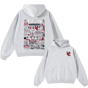 Puede incluir: Una sudadera con capucha de color gris claro con un bolsillo delantero y una capucha. La parte trasera de la sudadera con capucha presenta un gr&aacute;fico detallado con el texto "UNIVERSITY CUSTOM" y varias ilustraciones en rojo, blanco y negro. La parte delantera tiene un peque&ntilde;o logotipo "UC".