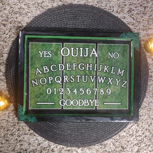 May include: A rectangular Ouija board with a black frame and green accents. The board features the words "YES", "OUIJA", and "NO", along with the alphabet, numbers, and the word "GOODBYE". The board is set on a dark gray surface.