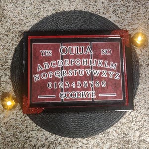 May include: A rectangular Ouija board with a black frame and a red and white patterned surface. The board features the alphabet, numbers, and the words "YES", "NO", and "GOODBYE". Decorative corners and two lit candles are also visible.