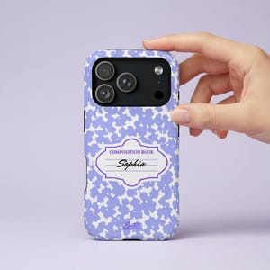 May include: A phone case featuring a light purple and white cow print pattern. In the center, a white label with a purple border displays the text COMPOSITION BOOK and the name Sophia in black script. The case has a black camera cutout area.