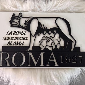 May include: A black and white wooden sign depicting the Capitoline Wolf with Romulus and Remus. The sign includes the text "LA ROMA NON SI DISCUTE SI AMA" and "ROMA 1927".