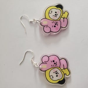May include: A pair of dangle earrings featuring a cartoon character design. Each earring has a silver-colored hook and a charm with a pink and yellow character. The character has a black outline and facial features.
