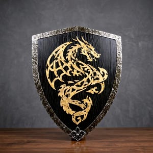 May include: A heater-style shield featuring a black wood-grain textured surface with a central gold-coloured dragon emblem. The shield is bordered by a hammered silver-toned metal rim with visible rivets and a decorative fleur-de-lis at the base.