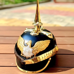 May include: A black military helmet with gold accents, including a prominent spike on top and an eagle emblem. The helmet features a gold band around the base and is displayed on a wooden surface.