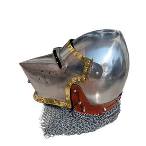 May include: A polished steel helmet with a pointed visor and brass detailing. The helmet has a leather neck guard and a chainmail lower edge. This medieval-style helmet is suitable for historical reenactment or display.