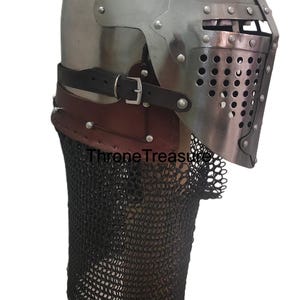Pu&ograve; includere: Un elmo a bacinetto in acciaio lucido con visiera incernierata e piastra frontale perforata, cinturino in pelle marrone con fibbia in metallo e una ventaglia di maglia metallica scura fissata alla base. Il testo ThroneTreasure &egrave; visibile.