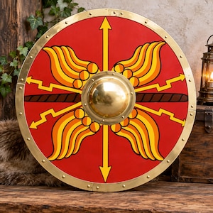 May include: A circular red shield featuring a central brass boss, surrounded by four yellow stylized wing motifs and four yellow lightning bolts. The shield is framed by a polished brass rim secured with evenly spaced brass rivets around the edge.
