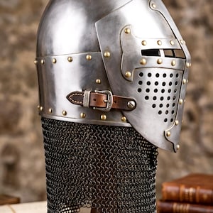 Pu&ograve; includere: Un elmo in stile medievale in acciaio lucido con visiera incernierata dotata di piastra frontale traforata, rivetti in ottone e un cinturino in pelle marrone con fibbia in metallo. Un camaglio in maglia metallica scura pende dalla base.