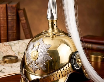 May include: A polished brass military helmet featuring a tall silver-toned spike topped with a white horsehair plume. The front displays a silver eagle emblem with a gold center. A brown leather chin strap with metal scales is attached to the sides.