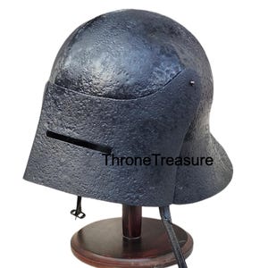 Op de afbeelding: Een donkergrijze metalen helm met vizier, tentoongesteld op een houten standaard. De helm heeft een getextureerd oppervlak en de tekst "Throne Treasure" is zichtbaar op het vizier.