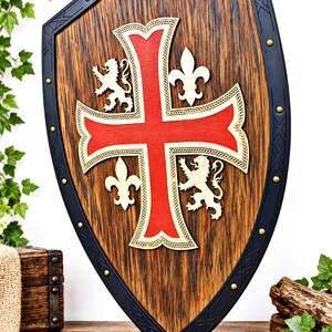 May include: A heater-style shield featuring a dark wood grain center with a prominent red cross outlined in gold. The shield includes two gold lion figures and two gold fleur-de-lis symbols. A black metal border with gold rivets frames the shield.