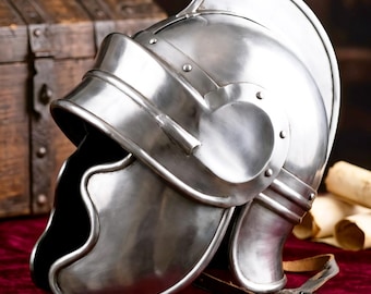 May include: A polished silver-colored metal helmet featuring a prominent central crest, a curved visor, and side cheek guards. The helmet includes a brown leather chin strap with a metal buckle attached to the lower side, showcasing a smooth finish.