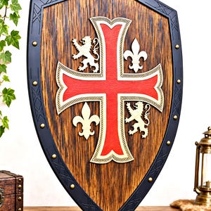 May include: A wooden shield featuring a large red cross at the center, flanked by two white lions and two white fleur-de-lis symbols. The shield has a dark, textured wood grain finish and a black border accented with small brass-colored studs.