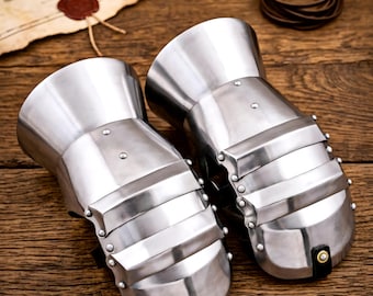 May include: A pair of polished steel gauntlets featuring articulated metal plates, a flared cuff, and small rivets. Each gauntlet includes a black leather strap with a central brass-colored stud at the fingertip area, designed for hand protection.