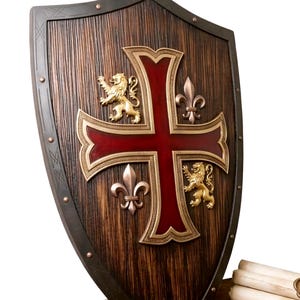May include: A pointed shield with a dark brown wood-grain finish, featuring a large central cross in deep red with gold-toned trim. The shield displays two gold-toned rampant lions and two metallic fleur-de-lis emblems, secured by small rivets.