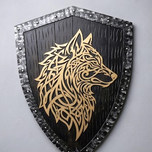Peut inclure: Un bouclier décoratif présentant un centre en bois noir avec un grain vertical et une silhouette de tête de loup dorée aux motifs de nœuds complexes. Le bouclier possède une bordure métallique argentée avec des rivets visibles sur le bord.