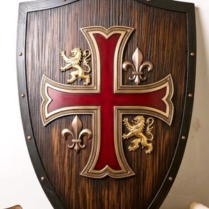May include: A wooden shield with a dark brown textured finish and a black metal border featuring copper rivets. A large red cross is centered on the shield, flanked by two gold-colored lion figures and two gold-colored fleur-de-lis emblems.