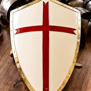 Könnte beinhalten: Ein weißes Schild mit einem roten Kreuz und einem goldfarbenen Rand. Das Schild ist von Metallhelmen und Rüstungsteilen umgeben, die alle auf einer Holzoberfläche liegen.