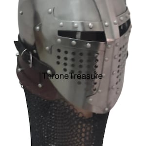 Pu&ograve; includere: Un elmo medievale in acciaio lucido con visiera a forma di T, fessure orizzontali per gli occhi e piastra frontale perforata. Un cinturino in pelle marrone scuro con fibbia in metallo fissa una ventaglia di maglia nera. Testo ThroneTreasure.