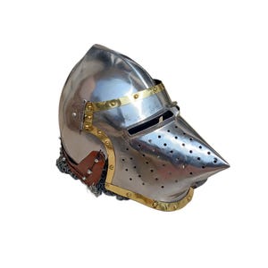 May include: A shiny steel helmet with a pointed visor and brass detailing. The helmet has small holes on the visor and a chainmail neck guard. The design is reminiscent of medieval armour.