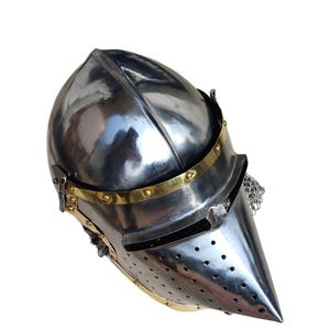 May include: A polished steel helmet with a pointed visor and brass detailing. The helmet has a riveted construction and a chainmail neck guard. The visor features small ventilation holes. This medieval-style helmet is suitable for historical reenactment or display.