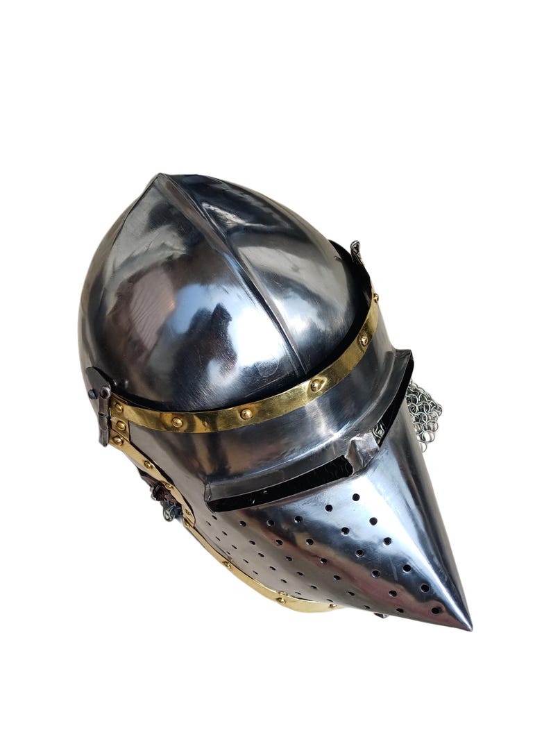 May include: A polished steel helmet with a pointed visor and brass detailing. The helmet has a riveted construction and a chainmail neck guard. The visor features small ventilation holes. This medieval-style helmet is suitable for historical reenactment or display.