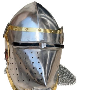 May include: A polished steel knight's helmet with brass detailing. The helmet has a pointed visor, ventilation holes, and a chainmail neck guard. The design is reminiscent of medieval armour.