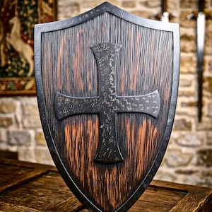 May include: A heater-style shield featuring a dark, textured wood grain finish with vertical streaks of brown and black. A large, centered cross with a distressed, dark metallic surface is mounted on the front, framed by a smooth, dark metal border.